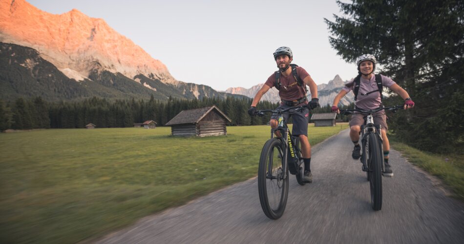 Zwei Radfahrer auf einem Weg, dahinter ein von der Abendsonne angestrahlter Berg | © Tiroler Zugspitz Arena/C. Jorda